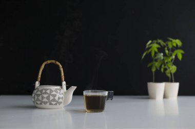 Çömlekçilikte marihuana bitkisi olan beyaz masa. Bardakta ve çaydanlıkta sıcak Cbd çayı.