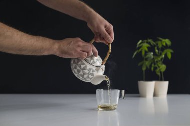 Çömlek içinde esrar bitki ile beyaz masa. Cam için sıcak CBD çayı dökmek el.