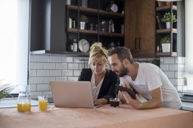 Mutfakta bir çift. Kadın dizüstü bilgisayar üzerinde çalışırken adam tablet üzerinde çalışıyor.