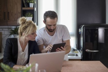 Mutfakta bir çift. Kadın dizüstü bilgisayar üzerinde çalışırken adam tablet üzerinde çalışıyor.