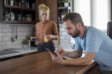 Mutfakta bir çift. Kadın içecekler getirirken erkek tablette tarama.