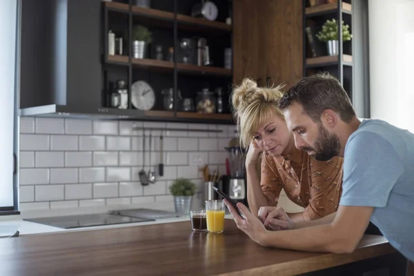 Couple in kitchen in the morning. They are browsing on tablet and smart phone.