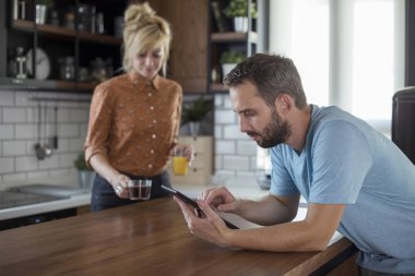 Mutfakta bir çift. Kadın içecekler getirirken erkek tablette tarama.