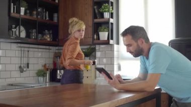 Mutfakta bir çift var. Karısı mutfak masasına meyve suyu ve kahve getiriyor kocası da tablet yazıyor.