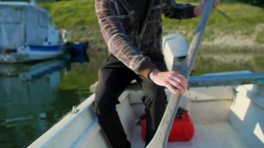 Beyaz orta yaşlı adam eski beyaz teknesinde kürek çekiyor. Gün doğumunda nehir marinasında. Bu balıkçı eski tahta küreği kullanıyor. Etrafında kayıklar ve yüzen evler ve arka planda ağaçlar var..