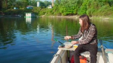 Balıkçı, yemi göle atıyor. Etrafında ağaçlar olan beyaz bir teknede. Adamın uzun kahverengi saçları var. İnşaat alanı arka planda. Mısır ve solucanlar su yüzeyine sıçrıyor damlalara neden oluyor..