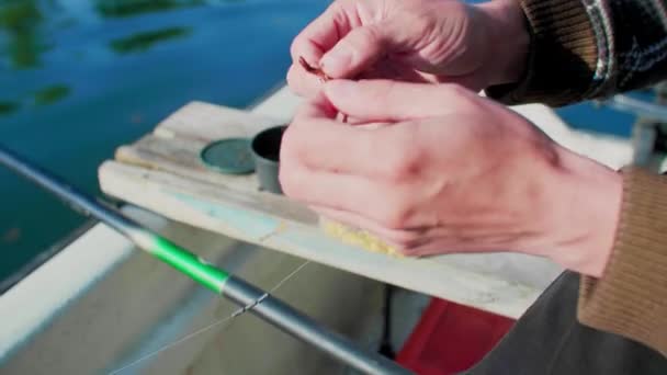 釣り針の餌としてワームを設定する手を閉じる 男は釣り竿を持っていて シンカーで並んでいる 川に浮かんでいる間 彼は船で働いている トウモロコシのボウルが背景にあります — ストック動画