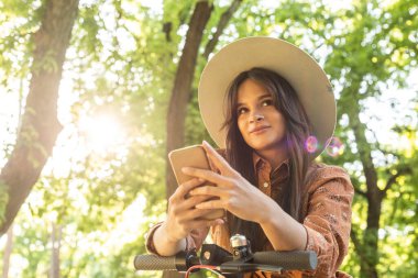 Telefonunda yazarken elektrikli scooterının yanında duran şapkalı güzel bir kız. Arka planda ağaçlar ve yeşil renkler baskın.