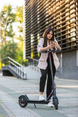 Telefonunda yazarken elektrikli scooter 'ın yanında duran genç ve güzel bir kız. Gülümsüyor, iş elbisesi giyiyor, arkasında modern mimari figürler var..