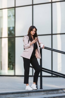 Genç ve güzel bir kız çite yaslanıyor ve cep telefonuyla yazarken gülümsüyor. Kahve içiyor, o da iş eğitimi almış, arka planda modern mimari var..