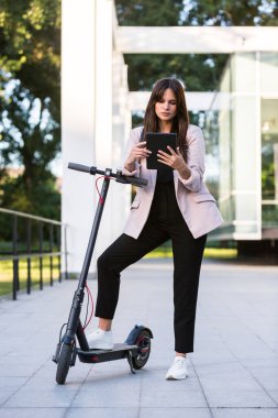Genç ve güzel bir kız, bir ayağı ayakta duran bir platformda, elektrikli scooterının yanında duruyor. Gülümsüyor, iş eğitimli, arka planda modern mimari var..