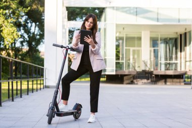 Genç ve güzel bir kız, bir ayağı ayakta duran bir platformda, elektrikli scooterının yanında duruyor. Gülümsüyor, iş eğitimli, arka planda modern mimari var..