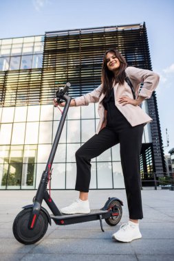Genç ve güzel bir kız telefonunda yazarken elektrikli scooterının yanında duruyor. Gülümsüyor, iş eğitimli, arka planda modern mimari var..