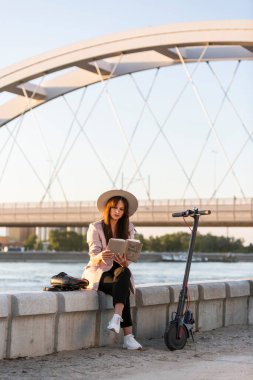 Elinde kitapla nehrin kenarında oturan şapkalı güzel bir kız var. Arkaplanda nehir ve köprü ağırlıklı iken, onun yanına bir elektrikli scooter park edilir..