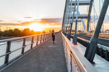 Genç ve güzel bir kız elektrikli scooterıyla köprüden geçiyor. İş eğitimi almış ve gülümsüyor. Arkaplan, modern bir köprünün parçalarından oluşuyor..