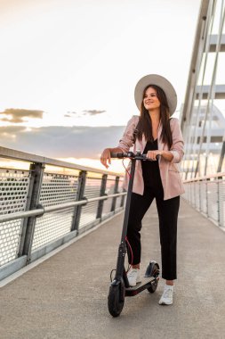 Genç ve güzel bir kız bir ayağı ayakta duran bir platformda elektrikli scooterının yanında duruyor. O bir şapka takıyor ve iş eğitimli. Arkaplan bir köprünün parçalarından oluşur