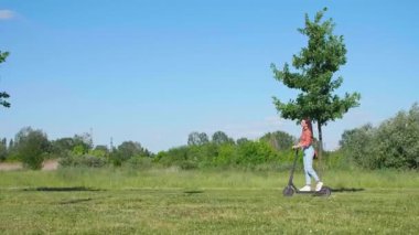 Genç ve güzel bir kadın parkın içinde elektrikli scooterla geziyor ve yeşil bir çevrede, bazı çekimlerde saçlarında rüzgar var..
