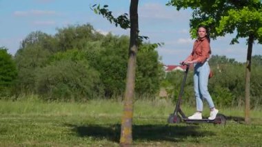 Genç ve güzel bir kadın elektrikli scooter ile çoğunlukla yeşil arka planda ve ağaçlarla dolu bir yerleşim yerinden geçiyor. Fotoğrafçıya el sallıyor..