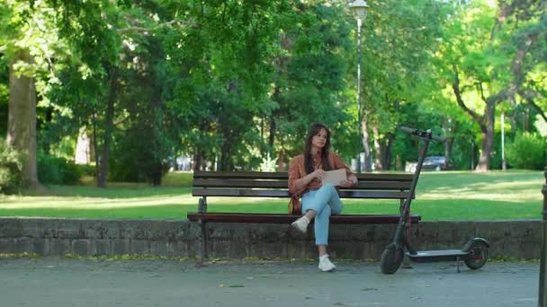 一个戴着帽子的年轻漂亮的女孩 手里拿着书 坐在公园的长椅上喝咖啡 她旁边停着一辆电动车 而后面主要是树木 — 图库视频影像