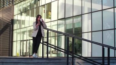 Genç ve güzel bir kadın çite yaslanıyor ve cep telefonuyla yazarken gülümsüyor. Kahve içiyor, o da iş eğitimi almış, arka planda modern mimari var..