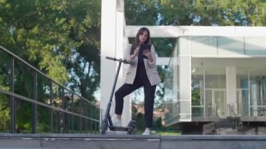 Genç ve güzel bir kadın elektrikli scooterının yanında ayakta duran bir ayakla duruyor. Gülümsüyor, iş eğitimli, arka planda modern mimari var..