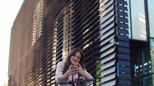 一个年轻漂亮的女人站在电动车旁边用她的手机 她面带微笑 穿着商务服装 以现代建筑为背景 — 图库视频影像