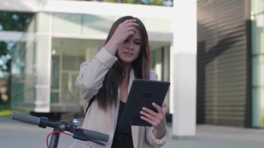 Genç ve güzel bir kadın elektrikli scooterının yanında ayakta duran bir ayakla duruyor. Gülümsüyor, iş eğitimli, arka planda modern mimari var..