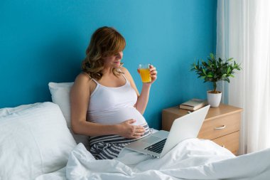 Dizüstü bilgisayarı üzerinde çalışırken yatağında oturan gülümseyen ve mutlu, hamile, beyaz bir kadın. Bir eli karnında ve meyve suyu içiyor.