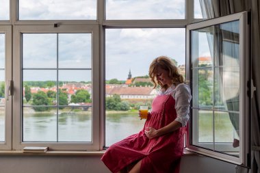 Eski kasabanın ve nehrin bir kısmı arka plandayken pencerenin kenarında oturan mutlu, hamile, beyaz kadın. Kollarını karnına doladığında bakışları aşağıya bakıyor..