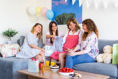 Bir grup gülümseyen arkadaş oturmuş, bebek partisini kutluyor ve hediyeleri açıyorlar. Atmosferik ve bir bebeğin beklentilerini kutluyorlar. Yer dekore edildi, aperatifler masada..