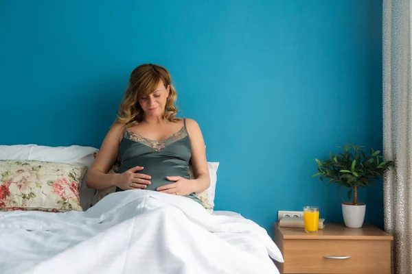 A smiling pregnant woman in a semi-sitting position in bed with her hands around her stomach. She's looking down at her stomach.