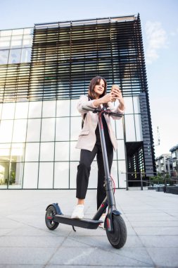 Genç ve güzel bir kadın telefonunda yazarken elektrikli scooterının yanında duruyor. Gülümsüyor, iş eğitimli, arka planda modern mimari var..