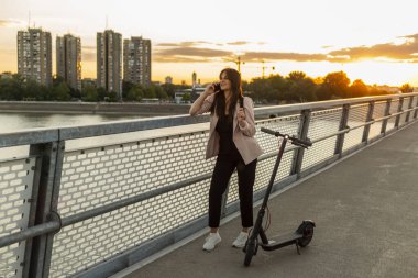 Genç, gülümseyen bir kadın telefonuyla konuşurken köprü çitinin yanında duruyor. Yanında elektrikli bir scooter park halindeyken, binalar, gün batımı ve arka planda köprü çiti ağırlıklı.