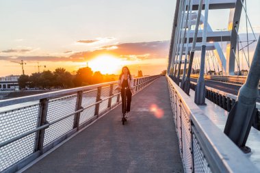 Genç ve güzel bir kadın elektrikli scooterıyla köprüden geçiyor. İş eğitimi almış ve gülümsüyor. Arkaplan, modern bir köprünün parçalarından oluşuyor..