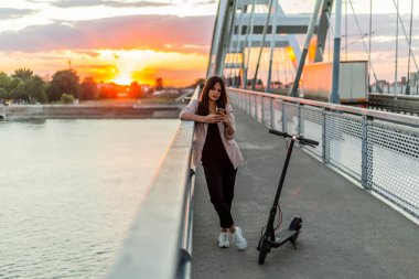 Genç, gülümseyen bir kadın köprü çitlerine yaslanıyor ve telefonuna yazı yazıyor. Onun yanına elektrikli bir scooter park edilmiş. Nehir, günbatımı ve arka planda modern bir köprünün parçaları yer alıyor..