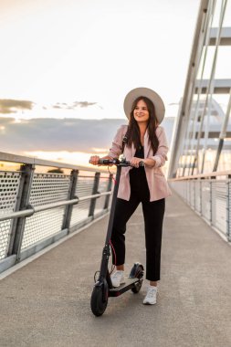 Genç ve güzel bir kadın bir ayağı ayakta duran bir platformda elektrikli scooterının yanında duruyor. O bir şapka takıyor ve iş eğitimli. Arkaplan bir köprünün parçalarından oluşur