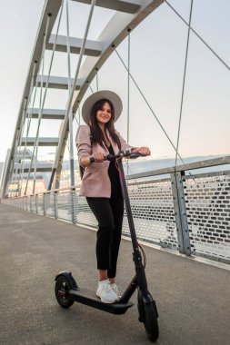 Genç ve güzel bir kadın bir ayağı ayakta duran bir platformda elektrikli scooterının yanında duruyor. O bir şapka takıyor ve iş eğitimli. Arkaplan bir köprünün parçalarından oluşur