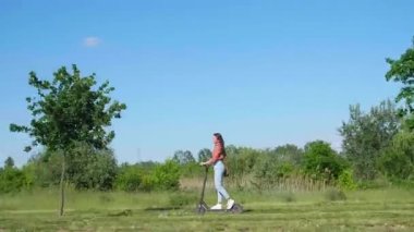 Genç ve güzel bir kadın parkın içinde elektrikli scooterla geziyor ve yeşil bir çevrede, bazı çekimlerde saçlarında rüzgar var..