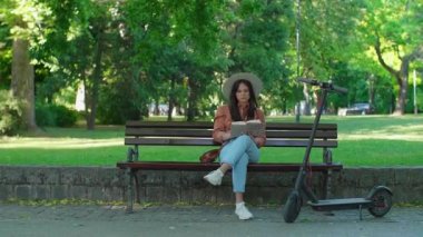 Şapkalı genç ve güzel bir kız kahve içerken bir bankta elinde kitapla oturuyor. Yanında elektrikli bir scooter park ederken, arka planda ağaçlar ağır basıyor..
