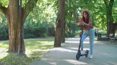Telefonunda yazarken elektrikli scooterının yanında duran şapkalı güzel bir kadın. Arka planda ağaçlar ve yeşil renkler baskın.