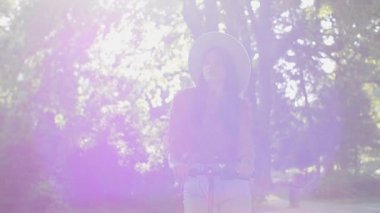 Şapkalı güzel bir kadın güneşli bir bahar gününü parkta geçirir ve elektrikli scooterına biner. Arka planda ağaçlar ağır basar..