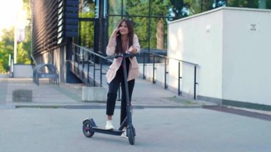 Telefonunu kullanırken elektrikli scooter 'ın yanında duran genç ve güzel bir kadın. Gülümsüyor, iş elbisesi giyiyor, arkasında modern mimari figürler var..