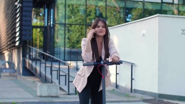 一个年轻漂亮的女人站在电动车旁边用她的手机 她面带微笑 穿着商务服装 以现代建筑为背景 — 图库视频影像