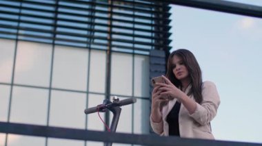 Genç ve güzel bir kadın telefonunda yazarken elektrikli scooterının yanında duruyor. Gülümsüyor, iş eğitimli, arka planda modern mimari var..