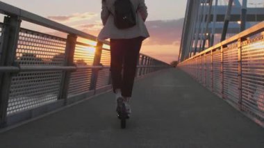Genç ve güzel bir kadın elektrikli scooterıyla köprüden geçiyor. İş eğitimi almış ve gülümsüyor. Arkaplan, modern bir köprünün parçalarından oluşuyor..