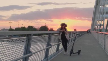 Genç, gülümseyen bir kadın köprü çitlerine yaslanıyor ve telefonuna yazı yazıyor. Onun yanına elektrikli bir scooter park edilmiş. Nehir, günbatımı ve arka planda modern bir köprünün parçaları yer alıyor..