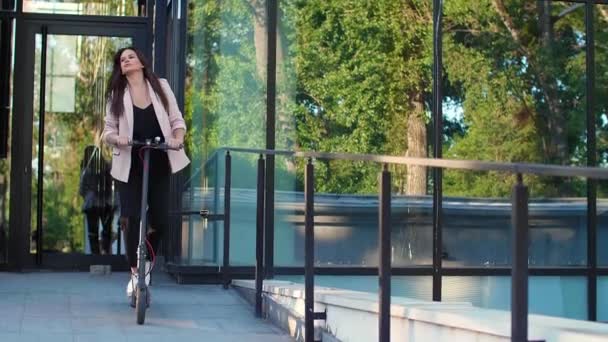 一个年轻漂亮的女人正在骑她的电动车 她面带微笑 穿着商务服装 以现代建筑为背景 — 图库视频影像