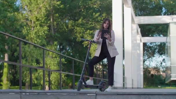 一位年轻漂亮的女士站在她的电动车旁边 一只脚踏在站台上 她面带微笑 受过商业训练 以现代建筑为背景 — 图库视频影像