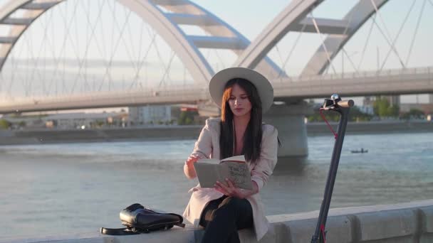 一个戴着帽子的年轻漂亮的女人手里拿着书坐在河边 一辆电动车停在她旁边 而身后主要是河流和一座桥 — 图库视频影像