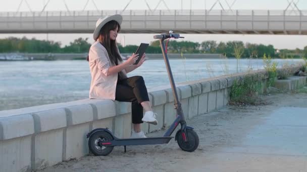 一个戴着帽子的年轻漂亮的女人手里拿着石碑坐在河边 一辆电动车停在她旁边 而身后主要是河流 太阳和一座桥 — 图库视频影像